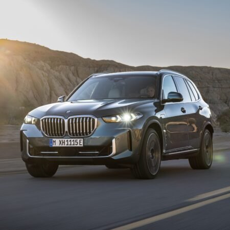 A BMW X5 Plug-in Hybrid electric vehicle driving through a desert landscape with the sun setting behind rocky hills.