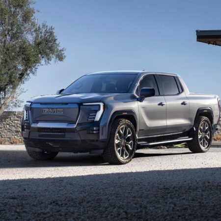 Side-front angle of the GMC Sierra EV Denali electric vehicle parked on a gravel driveway near a modern house