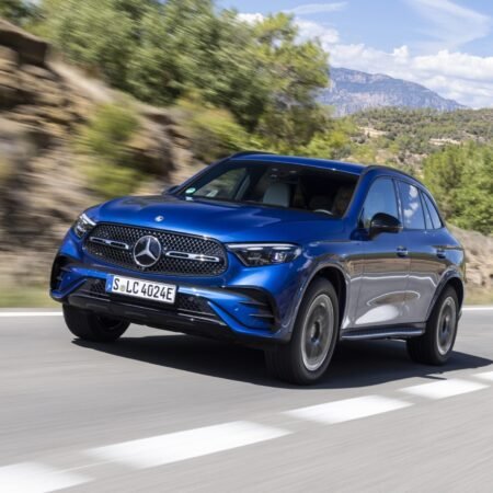 Blue 2025 Mercedes-Benz GLC 350e electric vehicle driving on a scenic road with hills and trees in the background