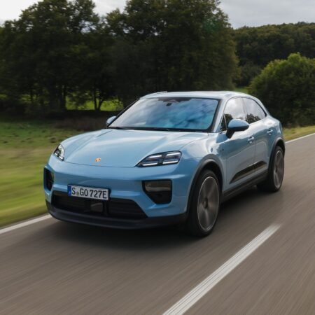 Light blue Porsche Macan EV electric vehicle captured from front-side angle while driving on a countryside road with green fields and trees